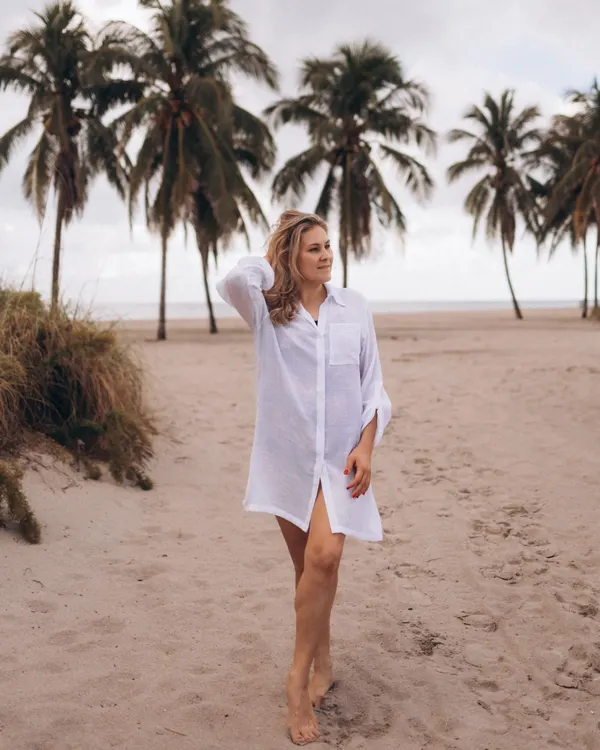 Olga, founder of Red Peony, walking on the beach in the white beach cover shirt, palm trees behind